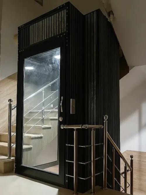 Compact home lift with black cabin beside a staircase.